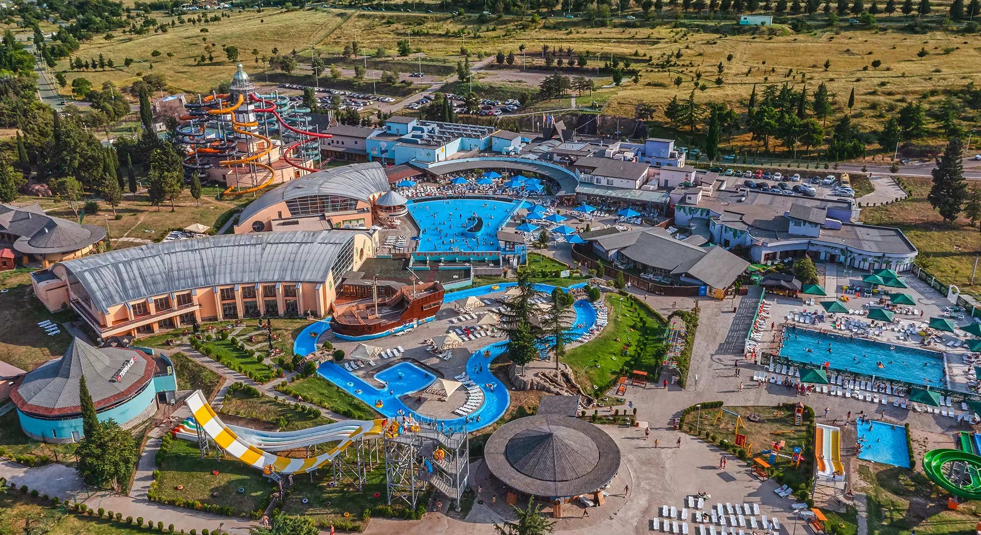 Gino Aquapark from above: the lighthouse slide tower, indoor halls, outdoor pools and a winding river spread across the park.